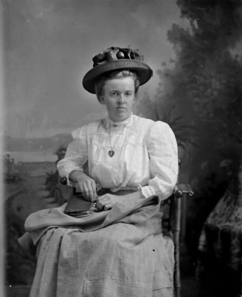 Studio portrait of an unidentified woman posing sitting. She has a purse and light coat on her lap, and is wearing a light-colored skirt and blouse, hat, and necklace with a heart-shaped locket.