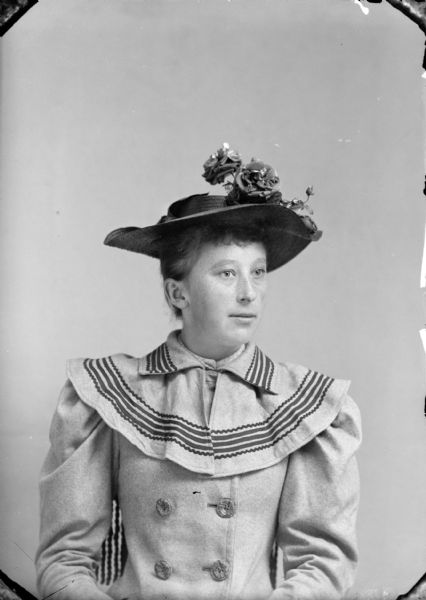 Studio portrait of an unidentified woman posing sitting. She is wearing a light-colored double-breasted coat with a wide collar and striped trim, and a dark-colored hat.