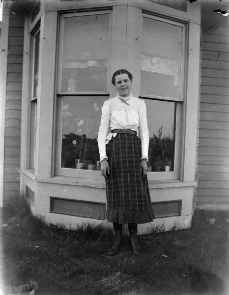 Exterior portrait of an unidentified woman posing standing in front of several windows of a wooden house. She is wearing a dark-colored plaid skirt and light-colored blouse.