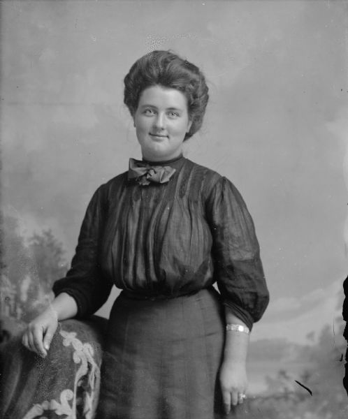 Three-quarter length studio portrait in front of a painted backdrop of an unidentified woman posing standing. She has her right hand resting on a cloth-covered chair on the left, and is wearing a dark-colored dress and a bracelet.