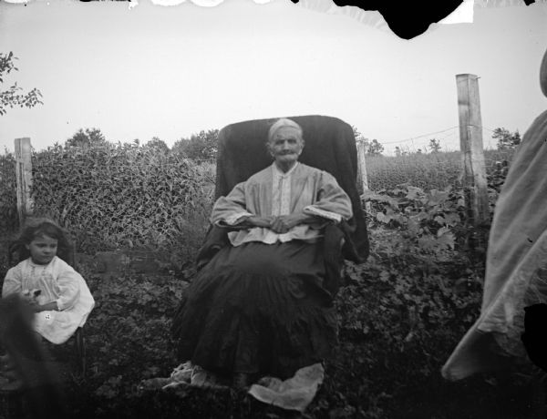 Outdoor portrait of an unidentified woman posing sitting in a cloth-covered chair in front of a wire fence. She is wearing a light-colored blouse and dark-colored skirt. On the left a girl is sitting in a small chair, wearing a light-colored dress and holding a kitten in her lap.