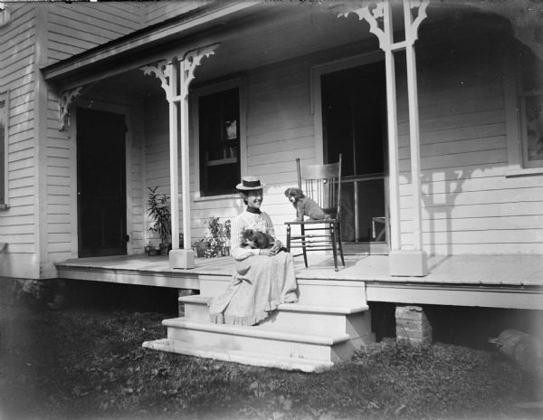 Outdoor portrait of an unidentified woman posing sitting on the porch steps of a wooden house. She is holding a cat on her lap, and on the porch is a puppy sitting on a wooden chair. The woman is wearing a light-colored skirt, blouse, and a straw hat.