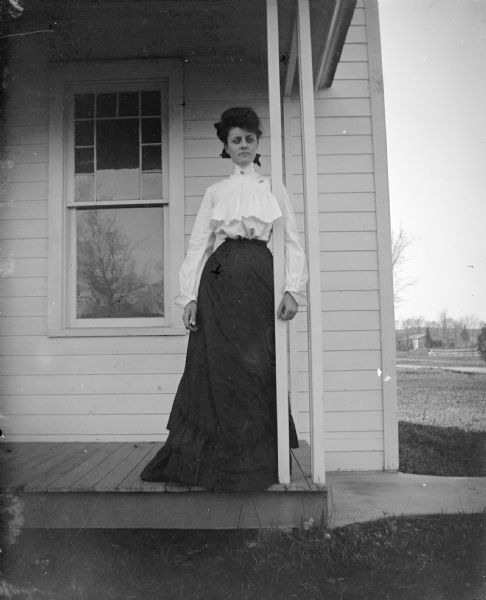Outdoor portrait of an unidentified woman posing standing and leaning against a narrow column of a wooden porch in front of a house. She is wearing a dark-colored skirt and light-colored blouse and collar pin.