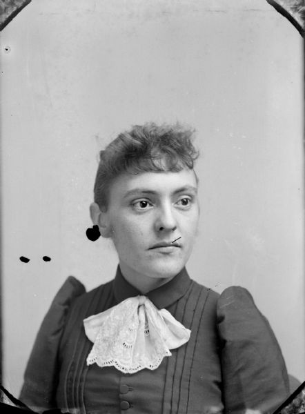 Quarter-length studio portrait of an unidentified woman posing sitting. She is wearing a dark-colored button-down and pleated blouse with a light-colored ruffle.