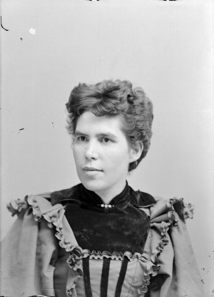 Quarter-length studio portrait of an unidentified woman posing sitting. She is wearing a light-colored dress with dark-colored velvet, ruffle trim, and a collar pin.
