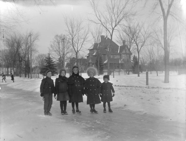 Outdoor group portrait of five children posing standing on ice, with a large house in the background. The house is identified as the Price house. Three of the children are wearing ice skates.