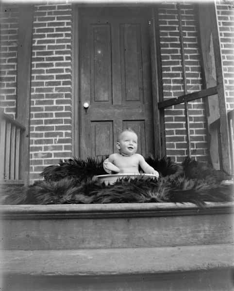 Exterior portrait of an unidentified infant posing sitting in a wash bowl on a dark-colored fur rug on a wooden porch in front of a brick building.