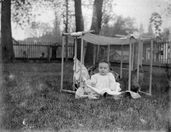 Outdoor portrait of an unidentified infant posing sitting on the ground underneath a makeshift awning set up in a yard. The infant is wearing a light-colored dress.