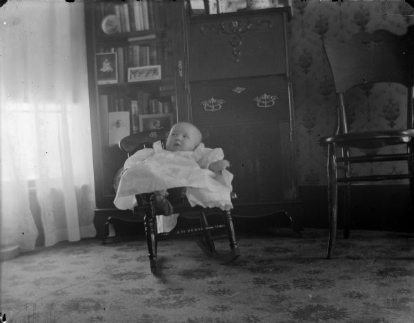 Indoor portrait of an unidentified infant posing sitting in a small wooden rocking chair in front of a bookshelf and secretary's desk. The infant is wearing a light-colored dress.