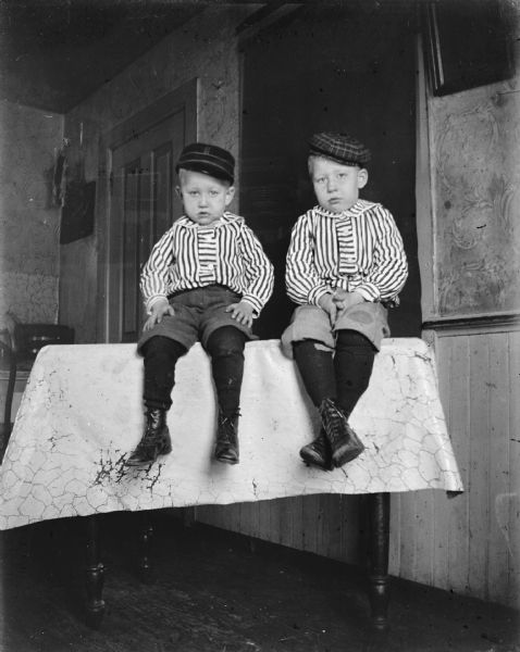 Indoor portrait of two European American boys posing sitting on top of a table. They are wearing short trousers, matching striped shirts, and caps.