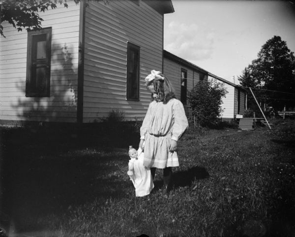 Outdoor portrait of an unidentified girl posing standing in a yard holding a doll. She is wearing a light-colored dress and a bow in her hair. In the background is a house.