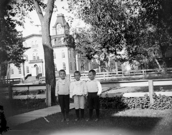 Outdoor portrait of three unidentified boys posing standing in a yard surrounded by a wooden fence, near a wooden walkway. The Jackson County building is in the background.