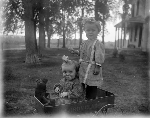 Exterior portrait of two unidentified girls posing standing and sitting in a children's wagon stenciled "Iron Clad." The girl standing on the right is holding flowers in her right hand, and the girl sitting on the left is sitting near a teddy bear. In the background is a house with a porch.