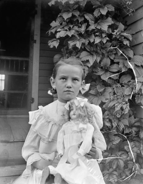 Outdoor portrait of an unidentified girl posing sitting on the wooden steps of a house near a climbing vine on a porch. She is wearing a dress and is holding a doll in her lap.