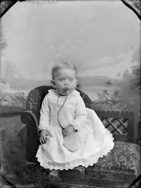 Studio portrait in front of a painted backdrop of an unidentified infant posing sitting on a chair. The child is wearing a light-colored dress and is holding/feeding from a bottle with a long nipple.