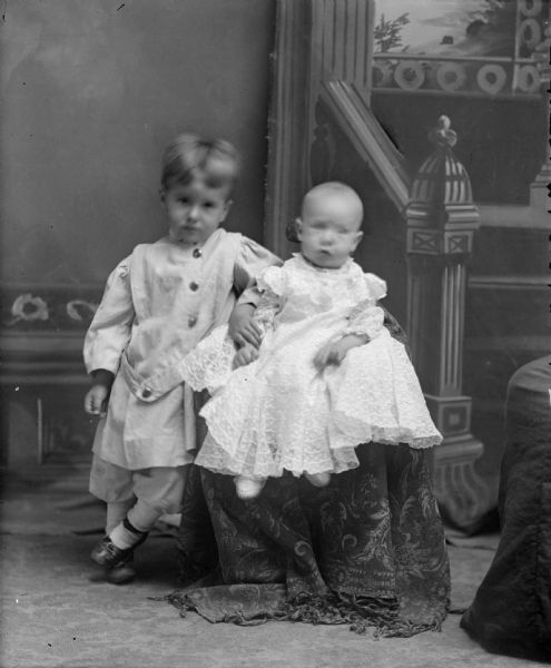 Indoor studio portrait in front of a painted backdrop of two unidentified children. One child is posing standing on the left, wearing a light-colored belted tunic and short trousers. The child next to him on the right is sitting up on a cloth-covered riser and is wearing a light-colored dress.