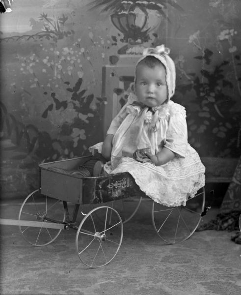 Studio portrait in front of a painted backdrop of an unidentified child posing sitting in a children's wagon. The child is wearing a light-colored dress and bonnet.
