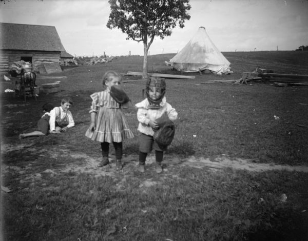 Outdoor group portrait of three children, two of them posing standing and one child lying down. One child is wearing what may be a catcher's mask, and the little girl is wearing a baseball mitt on her left hand. The group is in a field, and in the background is a tree, log cabin structure, and a light-colored tent.