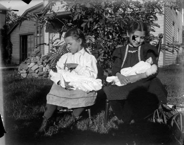 Outdoor portrait of two unidentified girls posing sitting in chairs on a lawn in front of a bush. The girls are holding dolls in their laps. On the far right is a baby carriage. In the background is a house.