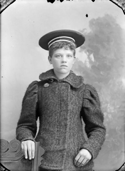 Waist-up studio portrait in front of a painted backdrop of European American boy posing. He has his right hand on the back of a wooden chair, and is wearing a dark-colored heavy winter coat and sailor-style hat.