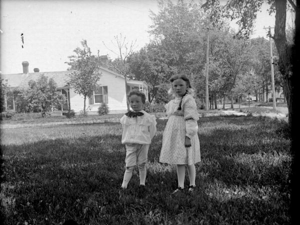 Exterior portrait of an unidentified boy and girl posing standing in a field in front of a wooden house. They are both wearing light-colored clothing.