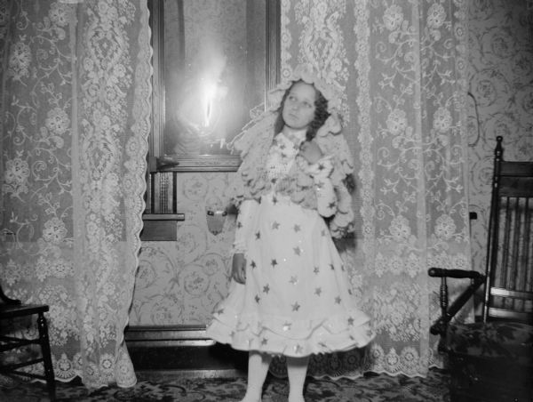 Indoor portrait of an unidentified girl posing standing in a room in front of lace curtains over windows. She is wearing a light-colored dress with a star-shaped print, and a light-colored shawl over her head. Photographer and flash are visible in the mirror in the center.