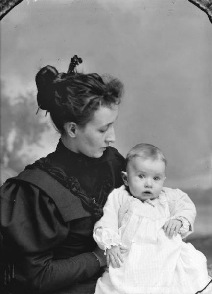 Waist-up studio portrait in front of a painted backdrop of an unidentified woman posing sitting and holding an infant in her lap. The woman is wearing a dark-colored dress and hair stays, and the infant is wearing a light-colored dress.