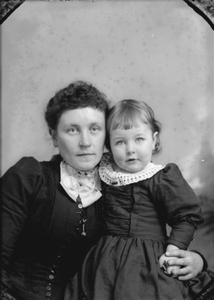 Waist-up studio portrait in front of a painted backdrop of an unidentified woman posing sitting. She is holding a girl in her lap who is posing standing. The woman is wearing a dark-colored dress with a button-down bodice, lapel pin, and light-colored neck bow. The girl is wearing a dark-colored dress and a wide light-colored lace collar.