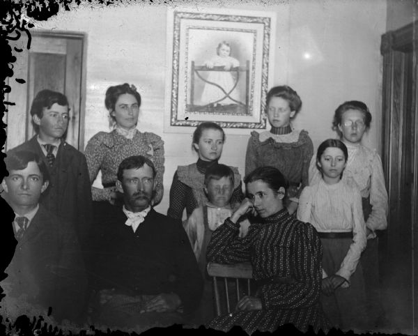 Indoor group portrait of nine unidentified individuals posing standing and sitting in front of a large studio portrait of a European American child wearing a light-colored dress. The boy in the center has his eyes tightly closed. The other people in the portrait, children and adults, are looking in several directions.