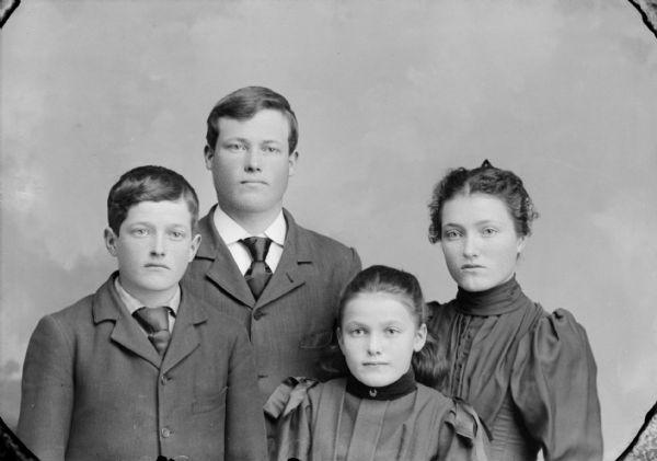 Waist-up studio group portrait in front of a painted backdrop of an unidentified man, woman, boy, and girl. The man and boy posing on the left are wearing dark-colored suit coats and neckties. The woman and girl posing on the right are wearing dark-colored dresses.