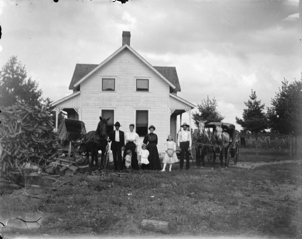 Outdoor group portrait of a family group posing standing in front of a wooden house and between two carriages drawn by horses. The group is identified as the O'Brien family.
