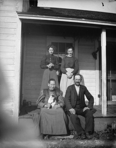 Outdoor group portrait of an unidentified group of three women and a man posing on the porch of a wooden house. The elderly man and woman are sitting on the porch edge, with the woman holding a cat in her lap.