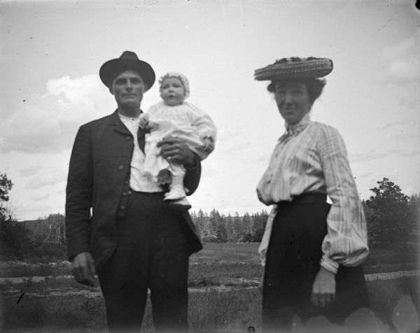 Outdoor portrait of an unidentified family group. The man is holding an infant in his arms, and near him on the right is a woman standing, and wearing a straw hat, dark-colored skirt, and light-colored blouse. The man is wears a dark-colored suit coat, trousers, and hat, and the infant is dressed in a light-colored dress and bonnet.