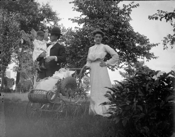 Outdoor group portrait of an unidentified family posing outdoors. On the left a man is holding a child on his hip. On the right, a woman is standing with one hand on her hip and the other hand on the handle of a wicker baby carriage, in which an infant is lying. They are posing among bushes and trees.