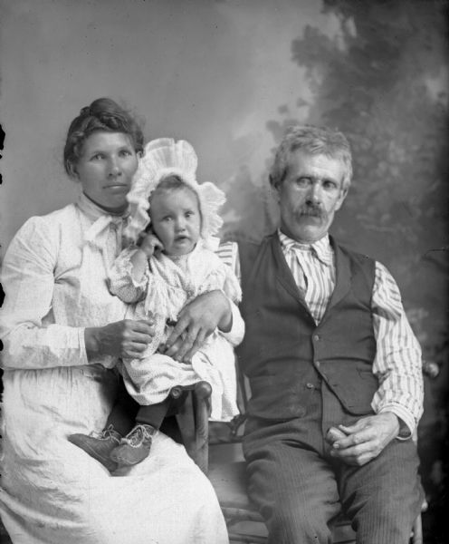 Studio group portrait in front of a painted backdrop of an unidentified family group posing sitting. The man on the right has a moustache and is holding a pipe in his lap. He is wearing a dark-colored vest and trousers, and a light-colored striped shirt. The woman on the left is holding a small child  in her lap. The woman and child are wearing light-colored dresses, and the latter a light-colored bonnet. 