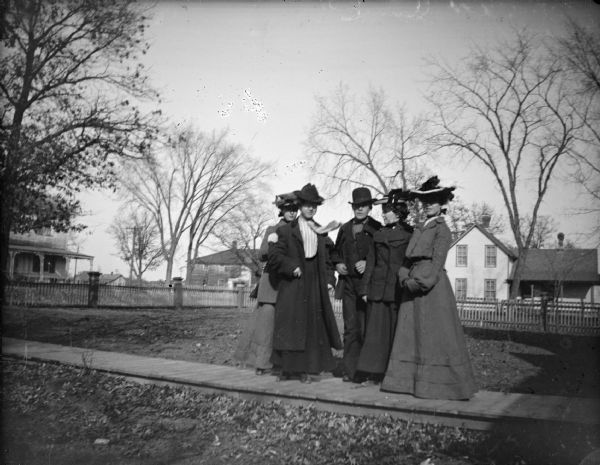 European American group of four women and a man posing standing on a wooden walkway in front several homes. Location identified as German Hill, in the Caves' front yard. The man is identified as probably Jim Kinley, and one of the woman on either side of him is probably his wife, Lizzie Caves Kinley.