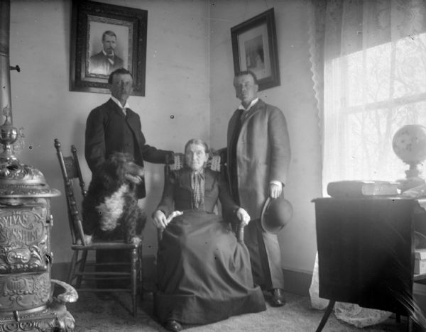 Indoor group portrait of an unidentified family group. There is a woman posing sitting in a chair between two men posing standing on either side of her. On the left near a wood-burning stove is a dog sitting on a chair.