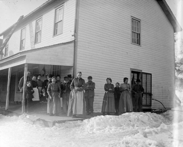 Outdoor group portrait of a large unidentified group of people posing standing on the porch and walkway of a wooden house. Snow is on the ground.