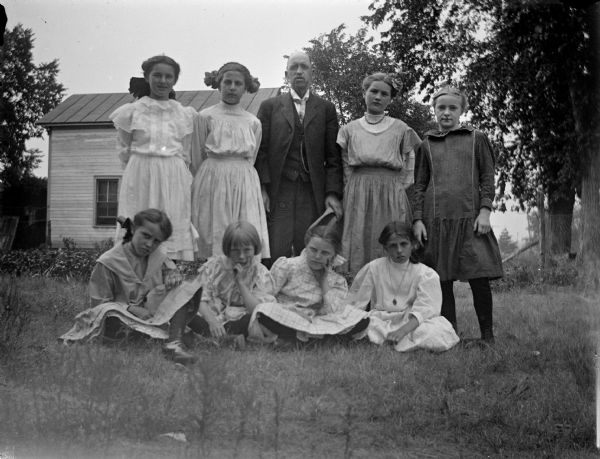 Outdoor group portrait of an unidentified group of people. Four young girls are sitting on the ground, and behind them are standing four girls, and one man posing in the center holding a hat. Behind them is a wooden house.