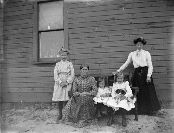 Outdoor group portrait of an unidentified group of people posing sitting and standing against the side of a house. A woman and two girls are sitting on a settee. The girl in the center is holding a doll, and the girl on the right has a dog in her lap. On the far left a young girl is standing, and on the far right a woman is standing with her hand on the back of the settee.