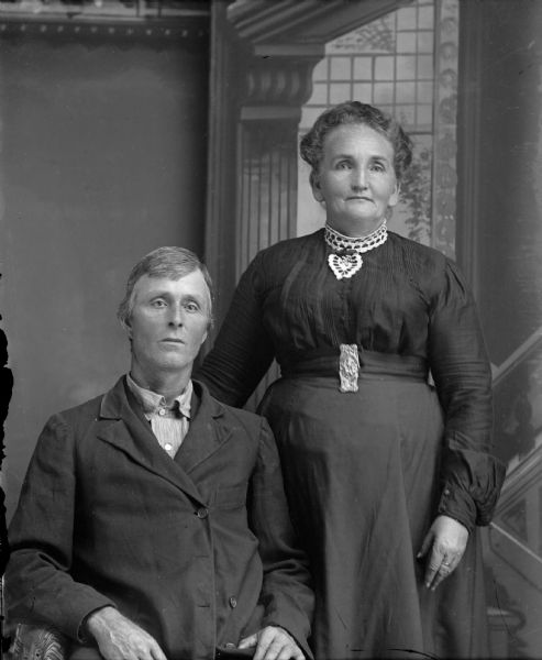 Studio portrait in front of a painted backdrop of an unidentified woman posing standing on the right next to a man sitting on the left. The woman is wearing a dark-colored dress with a light-colored collar. The man is wearing a dark-colored suit coat over a lighter-colored shirt.