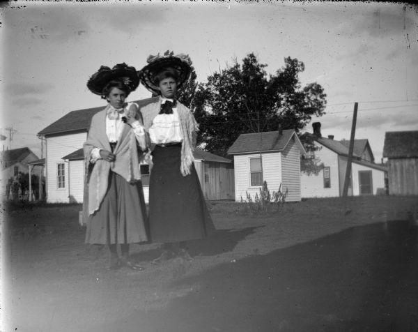 Outdoor portrait of two unidentified women posing standing. In the background are several wooden buildings. The women are wearing dark-colored skirts, light-colored blouses, shawls and large hats.