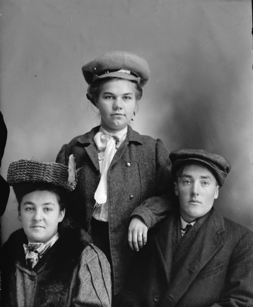 Studio group portrait of three unidentified people. In the center is a woman posing standing behind a woman sitting on the left, and a man posing sitting on the right. They are all wearing dark-colored coats and hats.