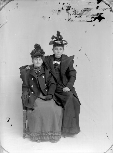 Full-length studio portrait of two young unidentified women posing sitting in front of a white backdrop. The woman on the left is sitting in the chair, and the woman on the right is sitting on the arm of the chair. They women are both wearing dark-colored dresses, long coats with puffy sleeves and wide lapels, and hats.