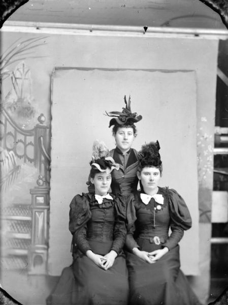 Studio group portrait in front of a painted backdrop of three unidentified women. In the center, one woman is posing standing behind two women posing sitting. They woman are all wearing dark-colored dresses and hats.