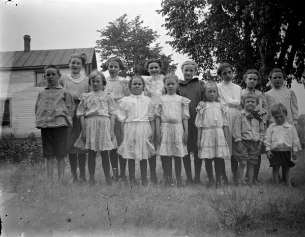 Outdoor portrait of a group of 14 unidentified children and young women posing standing in tall grass. There is a wooden building in the background on the left.