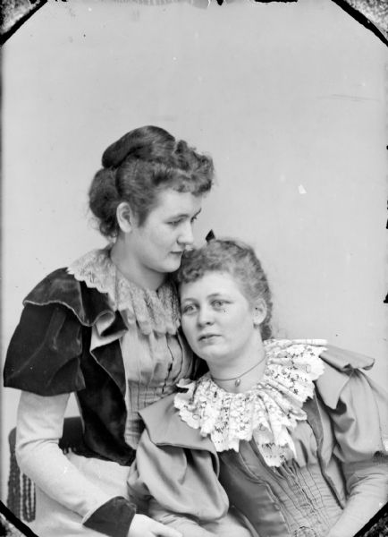 Studio portrait of two unidentified women. The woman on the right is leaning on the woman on the left. They are both wearing dresses with lace trim.