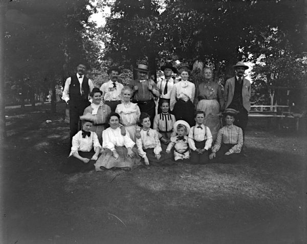Outdoor group portrait of a large, unidentified group of men, women, and children. They are posing sitting and standing in a park in front of trees and a picnic table.