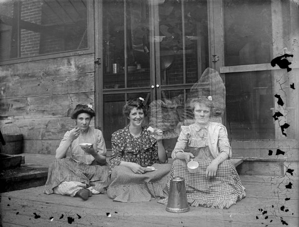 Double exposure. The dominant image is an outdoor portrait of three unidentified women posing sitting on a porch and eating. The fainter image is an outdoor portrait (sideways) of an unidentified girl posing sitting.
