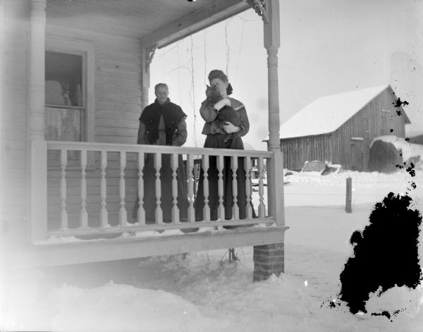 Outdoor portrait of two unidentified women posing standing on a porch on a wooden building. One of the women is holding a cat in her arms. Snow is on the ground, and in the background is a barn.